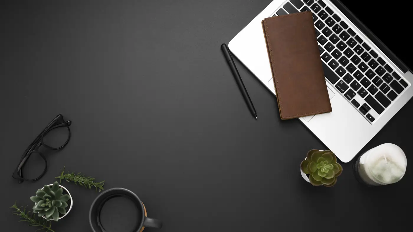 A laptop, a notebook, a pen and reading glasses on a dark desk.